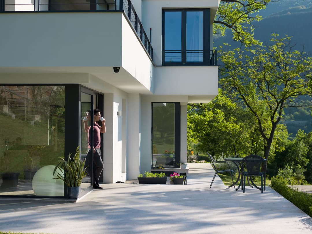 Modern house exterior with a person standing by the door, surrounded by greenery and outdoor seating.