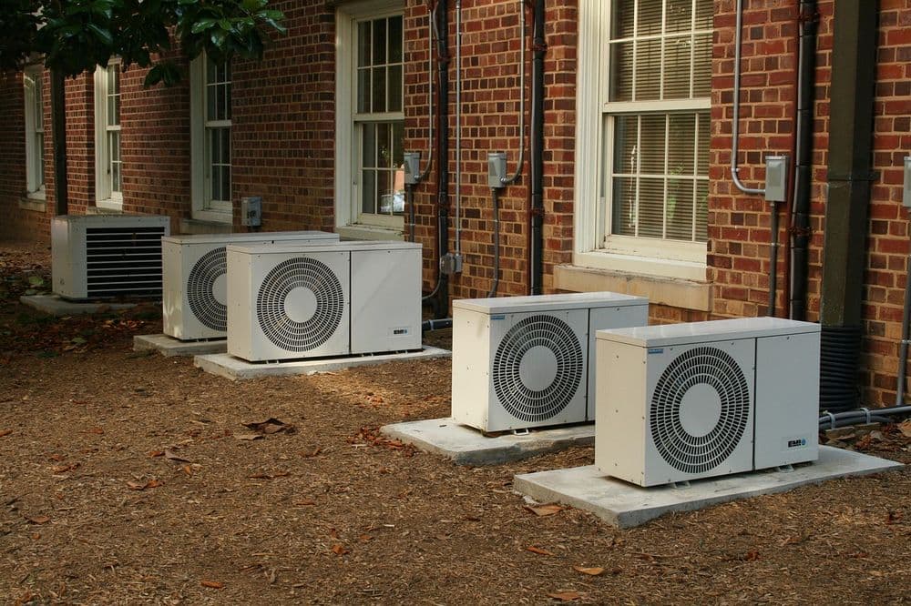 Four white air conditioning units installed on a building's exterior beside brick walls.
