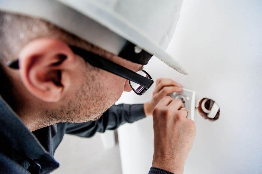 Electrician repairing wall outlet with tools, wearing safety gear and glasses.