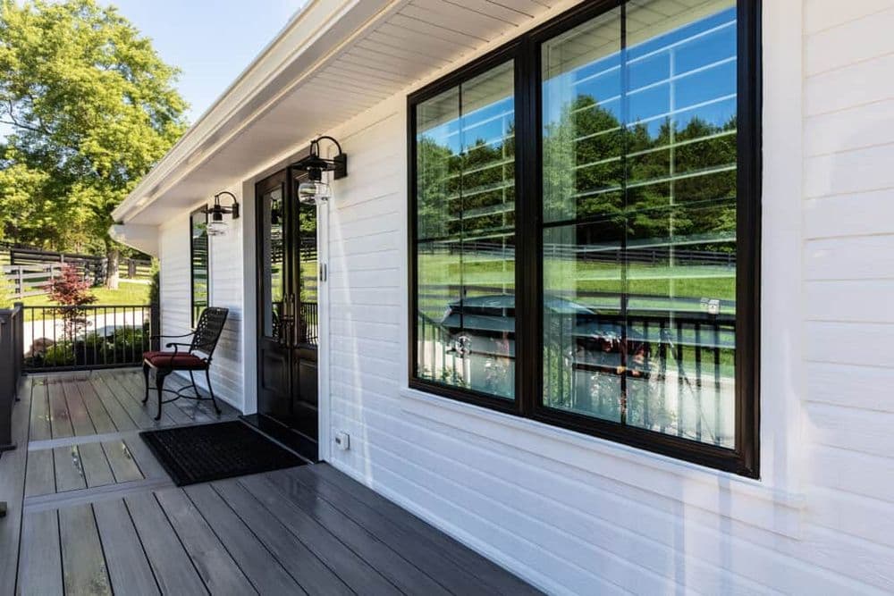 Modern home entrance with large windows reflecting greenery, featuring black door and porch seating.