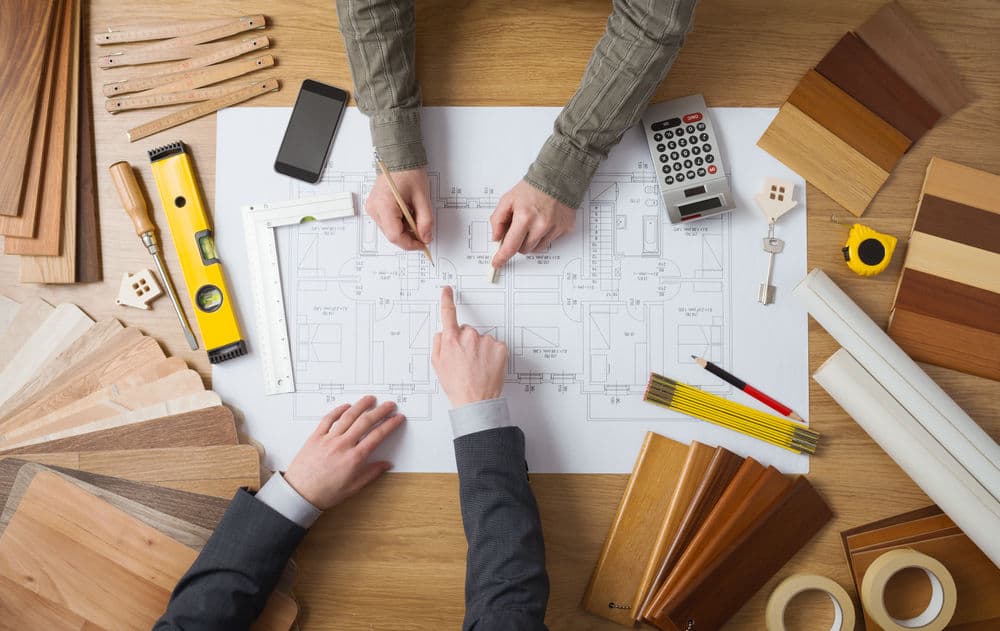 Hands analyzing architectural plans with tools and wood samples on a wooden desk.