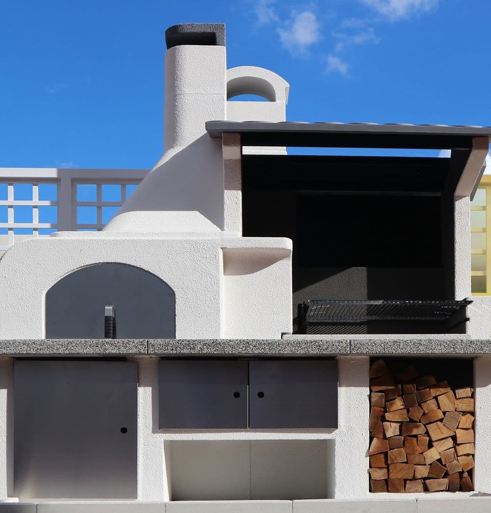 Modern outdoor kitchen with pizza oven, grill, and stacked firewood under a blue sky.