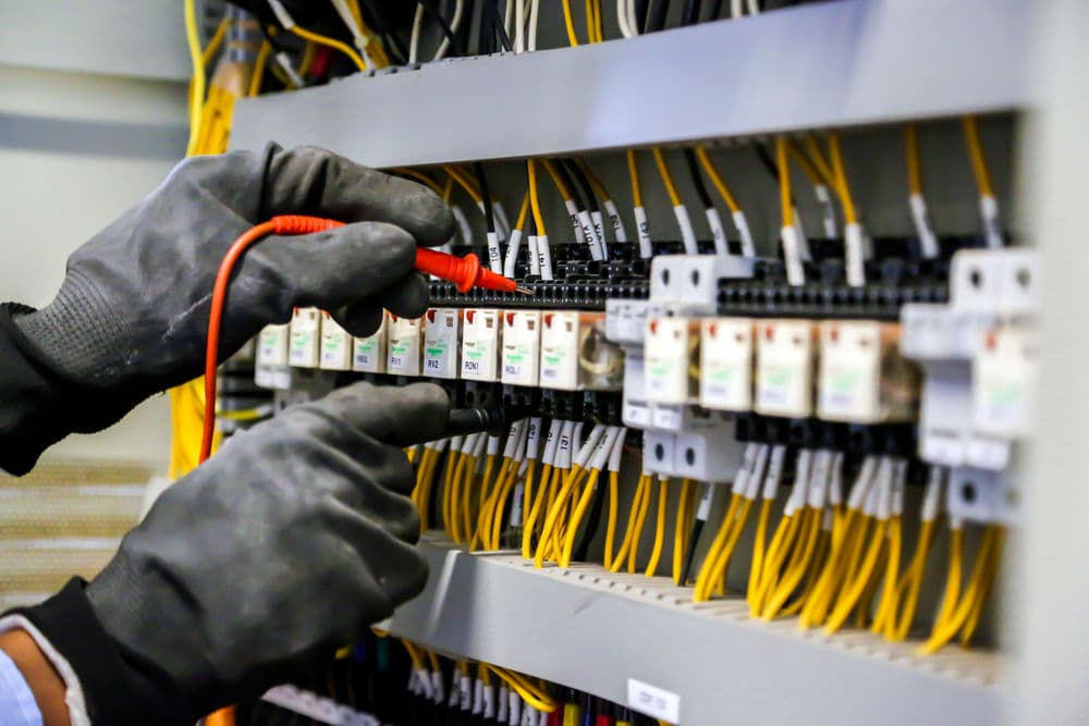 Electrician testing circuit breakers with multimeter in electrical panel. Safety gloves worn.