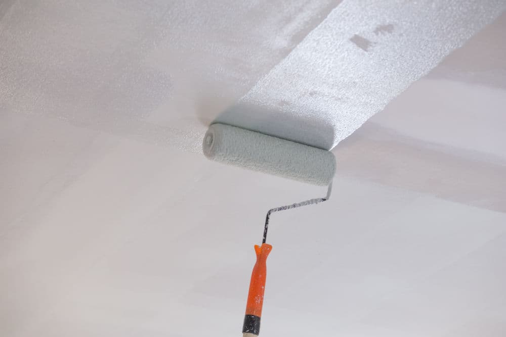 Paint roller applying light gray paint to a smooth, freshly prepared ceiling surface.